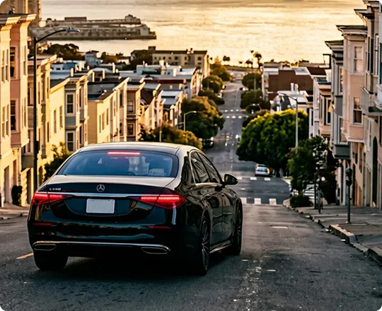 Rear view of a black Mercedes-Benz sedan driving down a steep San Francisco street lined with row houses toward the bay at sunset.