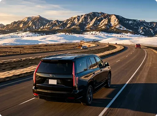 Rear view of a black Cadillac Escalade driving along a curving highway toward the snow-dusted Flatirons near Boulder at golden hour.