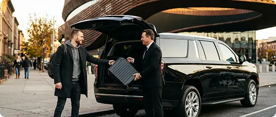 A chauffeur in a black suit loads a traveler’s suitcase into the trunk of a black luxury SUV on a Brooklyn street outside an arena.